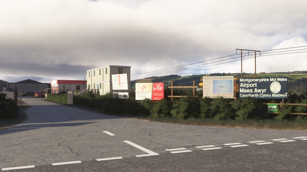 L'aéroport Welshpool de Burning Blue Design pour MSFS 2020 et MSFS 2024, en vue SIM de l'entrée principale de l'aéroport depuis la route locale avec des panneaux, des bâtiments et des lignes électriques.